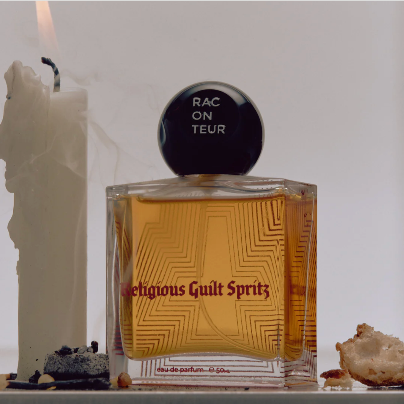 Perfume bottle labeled 'Religious Guilt Spritz' with a candle and bread crumbs on a white surface.